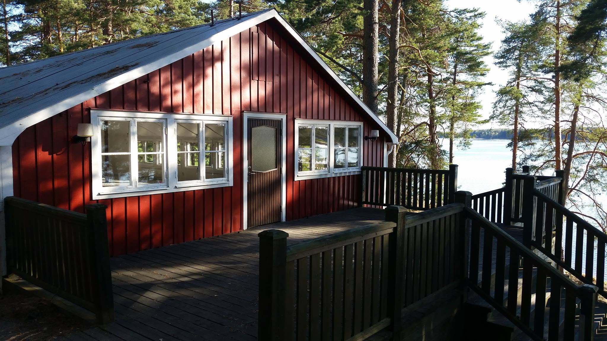 En röd trästuga med vitmålade fönster, omgiven av träd, med utsikt över en sjö i solskenet.