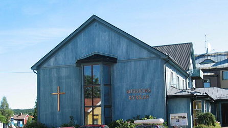 En blå träkyrka med ett kors och bilar parkerade utanför en solig dag.