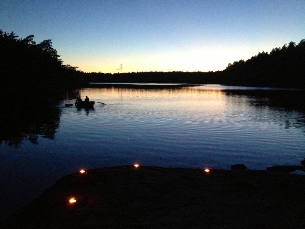 En kanot med två personer på en stilla sjö vid solnedgången, med tända ljus på den klippiga stranden i förgrunden.