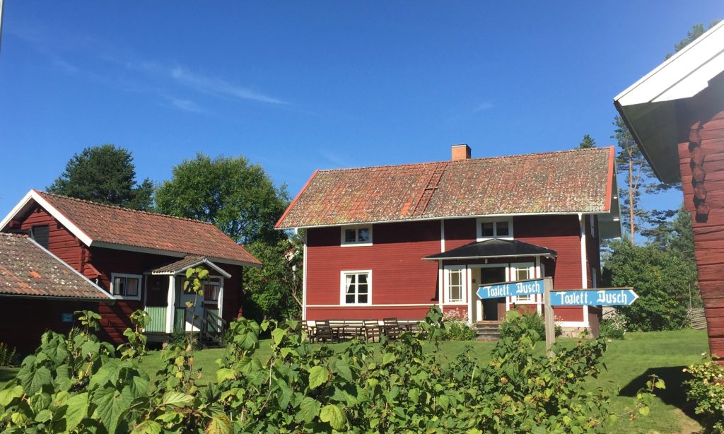 Röda trähus med tegeltak en solig dag, med blå skyltar där det står ”Toalett” och ”Dusch”.