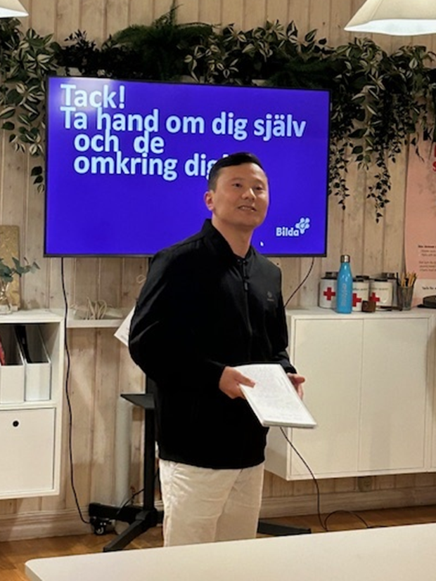 En person står och håller i några papper, med en skärm bakom sig där det visas svensk text och växter ovanför.