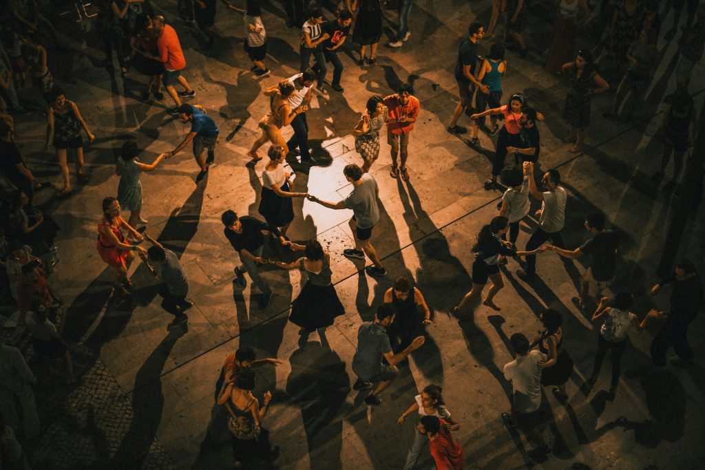 En grupp människor dansar tillsammans utomhus på kvällen och kastar långa skuggor på marken.