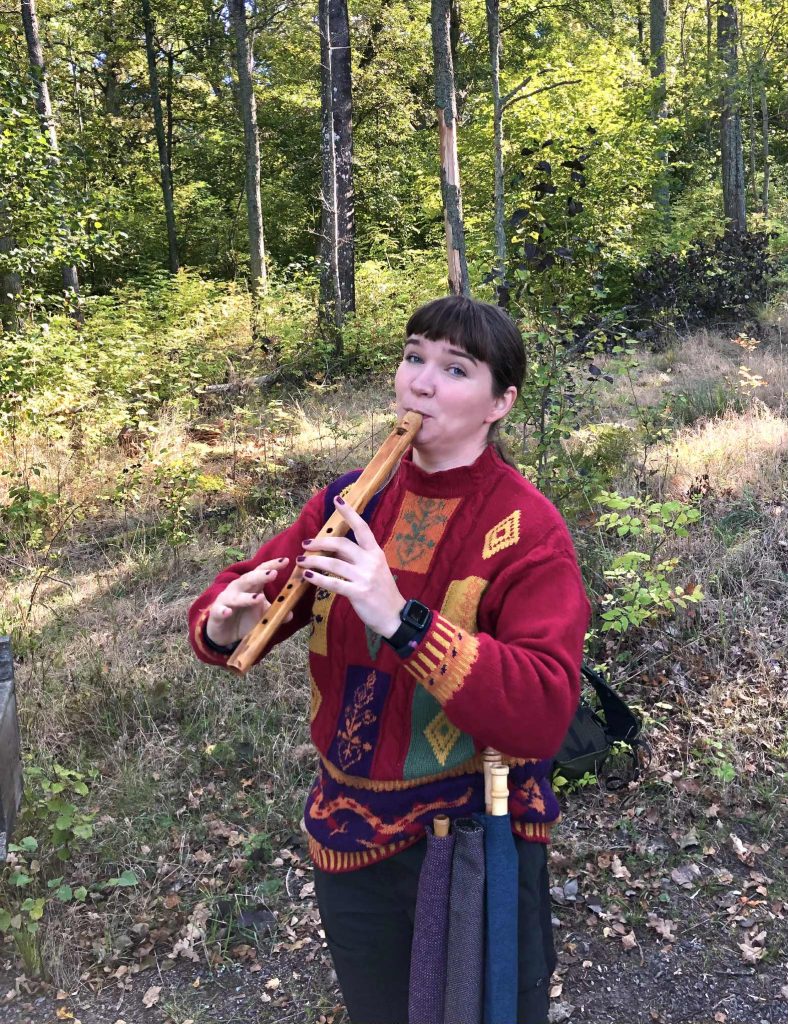 En kvinna i en färgglad tröja spelar på en träflöjt utomhus i en solbelyst skog.
