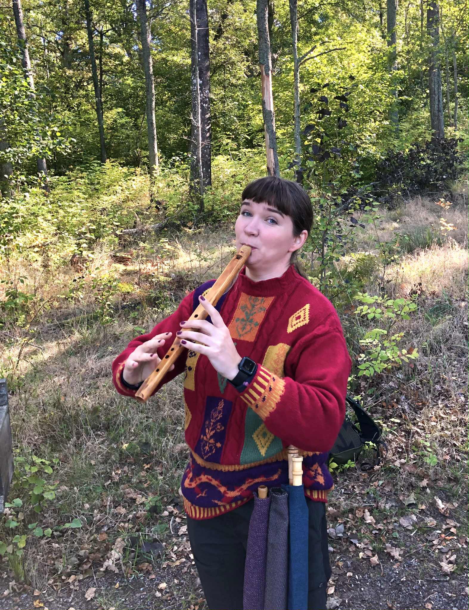 En kvinna i en färgglad tröja spelar på en träflöjt utomhus i en solbelyst skog.