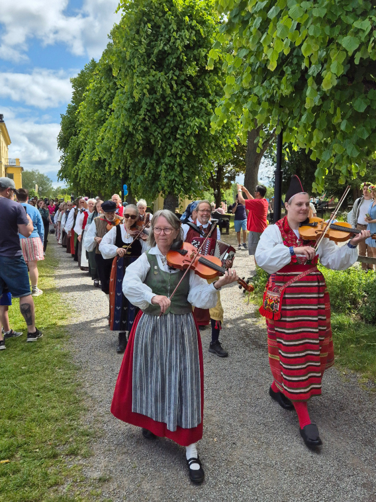Människor i traditionella kläder spelar fioler och leder en parad utomhus med åskådare och gröna träd.