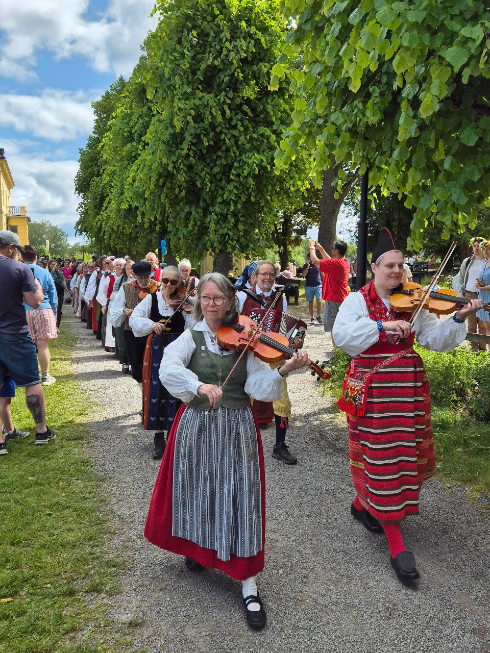 Människor i traditionella kläder spelar fioler och leder en parad utomhus med åskådare och gröna träd.