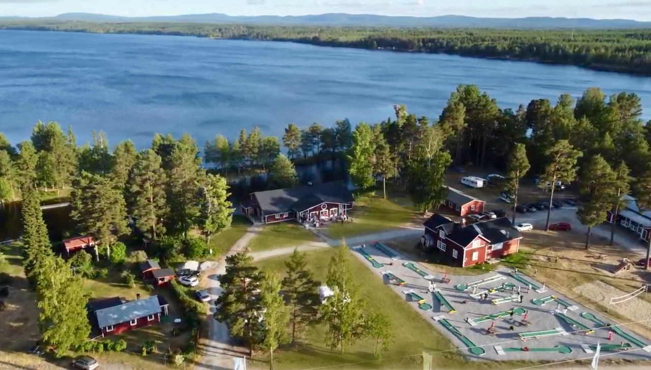 Flygfoto över stugor, träd och en minigolfbana vid en sjö nära en stor blå sjö.