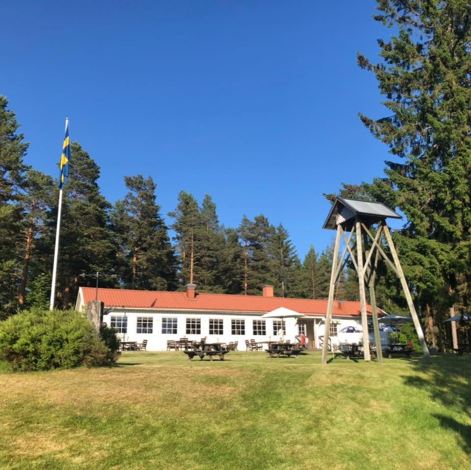 Vit byggnad med rött tak, picknickbord, hög flaggstång och klocktorn omgiven av träd under en blå himmel.