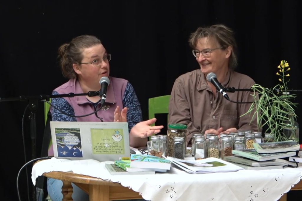 Två kvinnor sitter vid ett bord med mikrofoner, burkar, böcker och växter och pratar och ler under en diskussion.