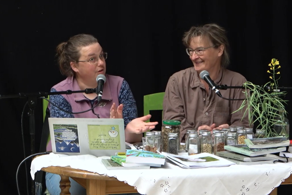 Två kvinnor sitter vid ett bord med mikrofoner, burkar, böcker och växter och pratar och ler under en diskussion.