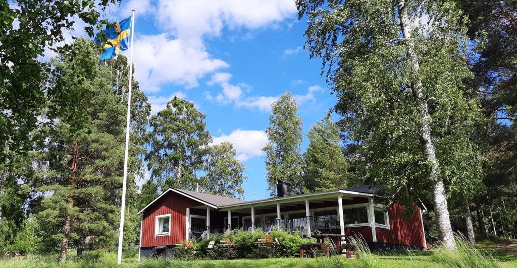Ett rött trähus med veranda, omgivet av träd, och en svensk flagga på en flaggstång under en blå himmel.