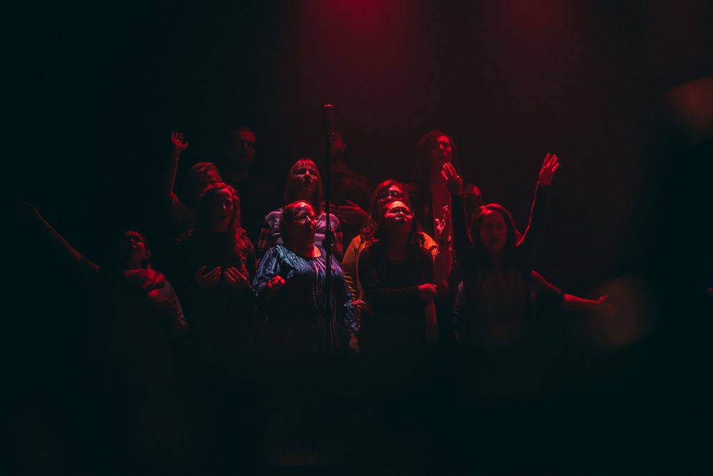 En grupp människor som sjunger på scenen i dramatisk röd belysning med uppsträckta armar.