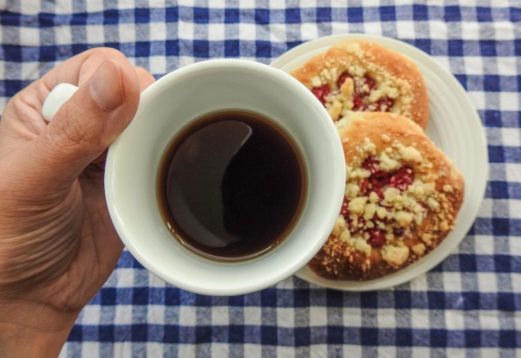 En hand som håller en kopp kaffe ovanför en tallrik med två bakverk på en blå- och vitrutig duk.