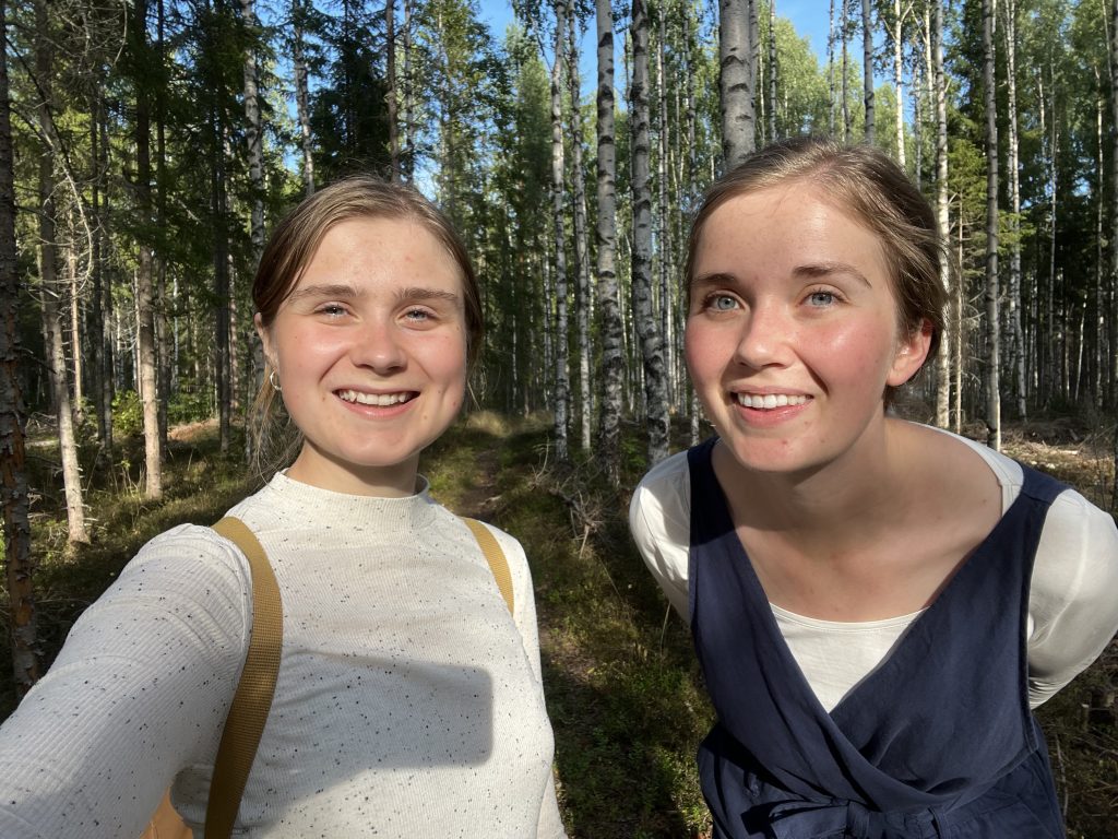 Två unga kvinnor ler i en solbelyst skog med höga träd och gröna bladverk i bakgrunden.