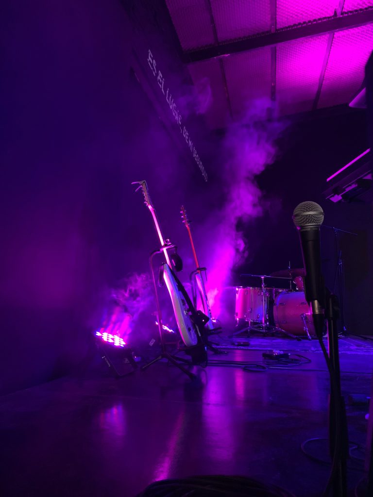Två gitarrer, ett trumset och en mikrofon på en scen med lila belysning och dimma.