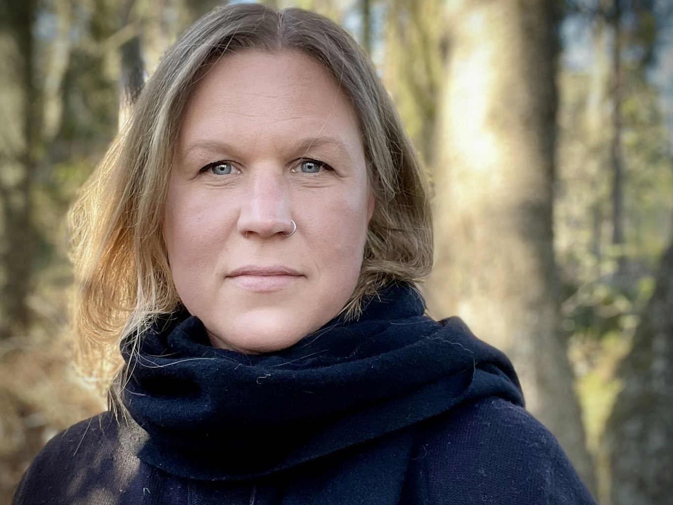 En kvinna med axellångt hår och en svart sjal står utomhus med träd i bakgrunden.