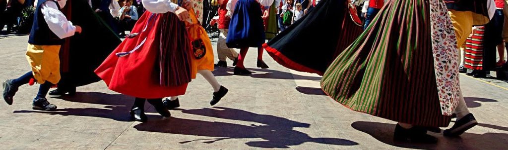 Människor som dansar utomhus i färgglada traditionella kläder, med virvlande kjolar som kastar skuggor på marken.