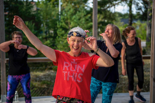 Fyra kvinnor dansar och ler utomhus; kvinnan längst fram har på sig en röd tröja med texten ”Run This” och ett hårband.