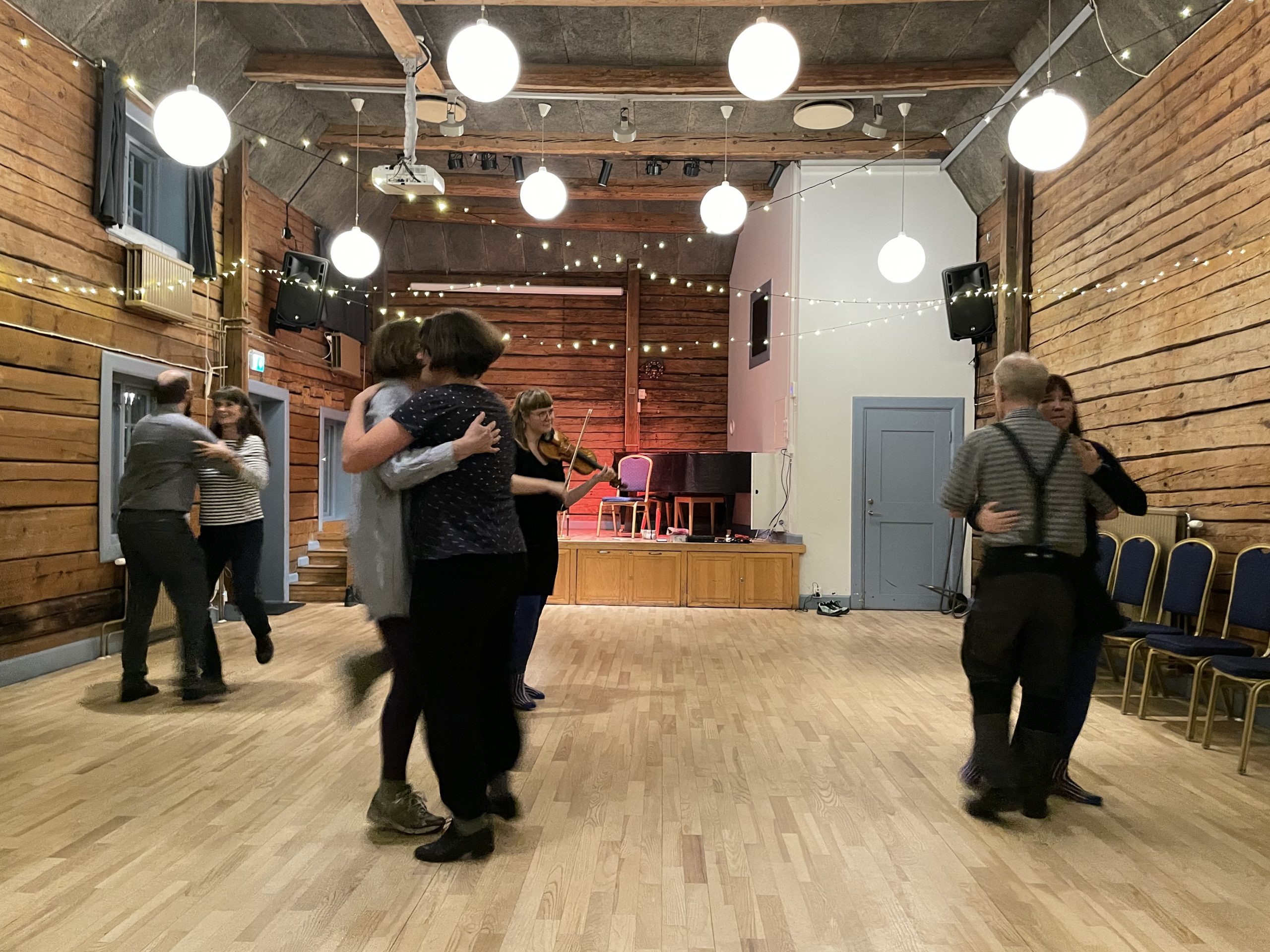 Människor som dansar i par i en träsal med ljusslingor och en musiker som spelar fiol på scenen.