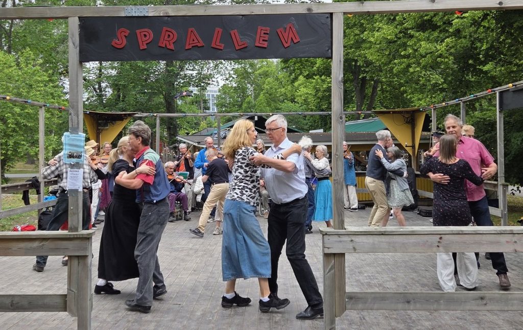 Människor som dansar i par utomhus under en skylt med texten ”Sprallen”, omgivna av träd och med musiker i bakgrunden.