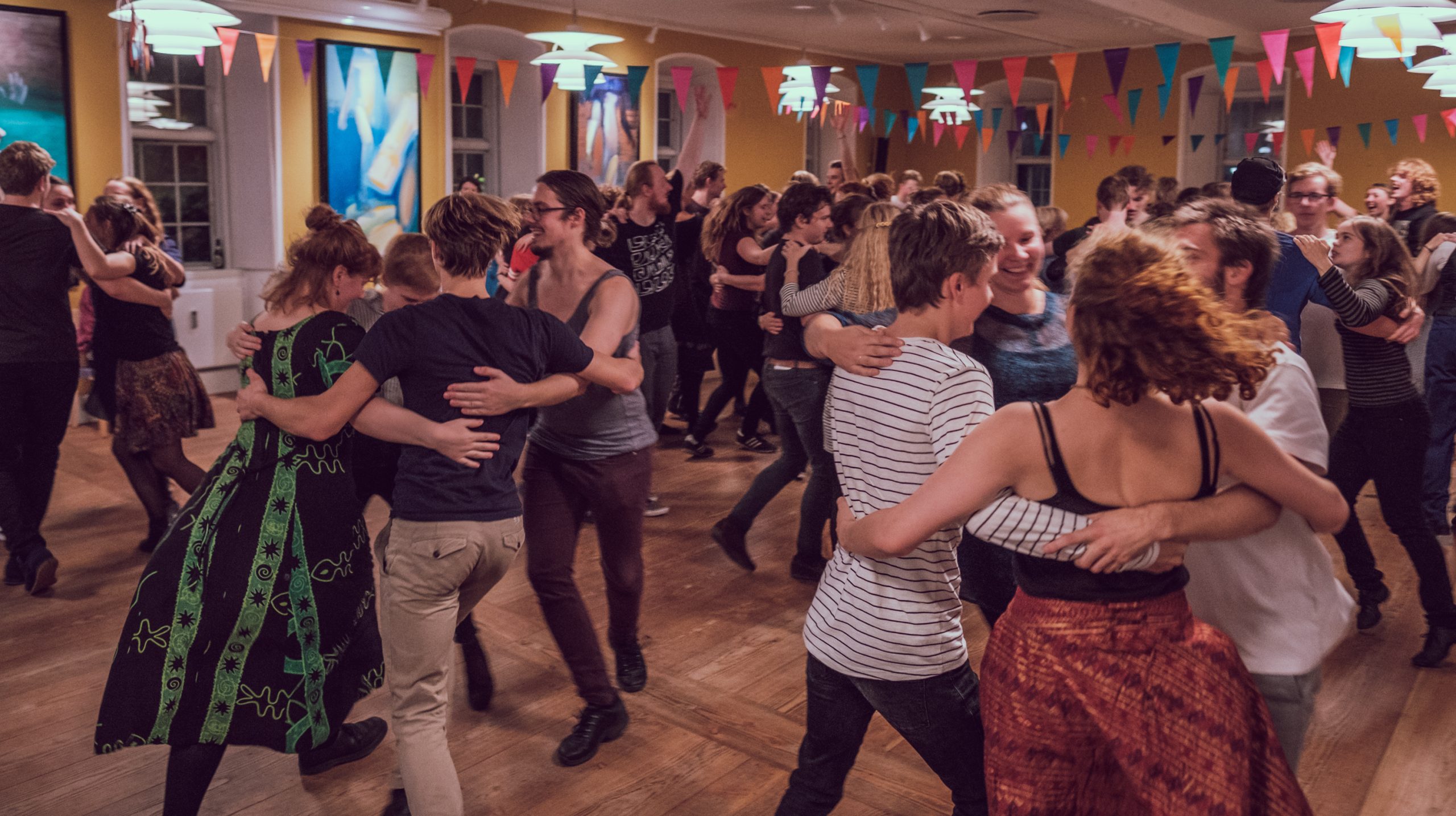 Människor som dansar i par på en livlig fest inomhus med färgglada flaggor och varm belysning.