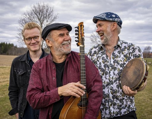Tre leende män står utomhus med musikinstrument en molnig dag.