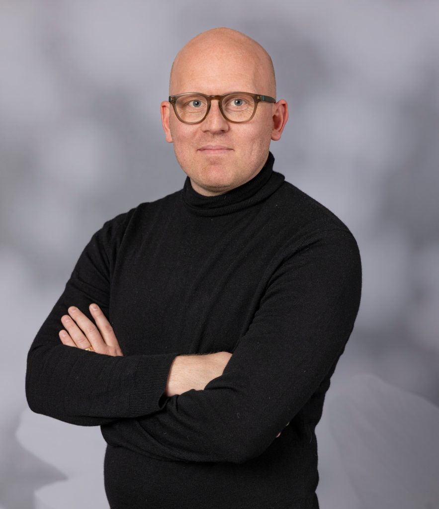 En person med glasögon och svart polotröja står med korsade armar mot en grå bakgrund.