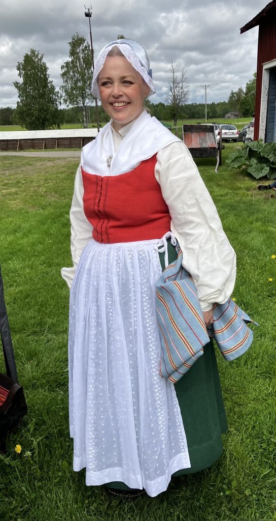 En kvinna som ler utomhus, klädd i traditionell folkdräkt med vitt förkläde och med randiga tygstycken i händerna.