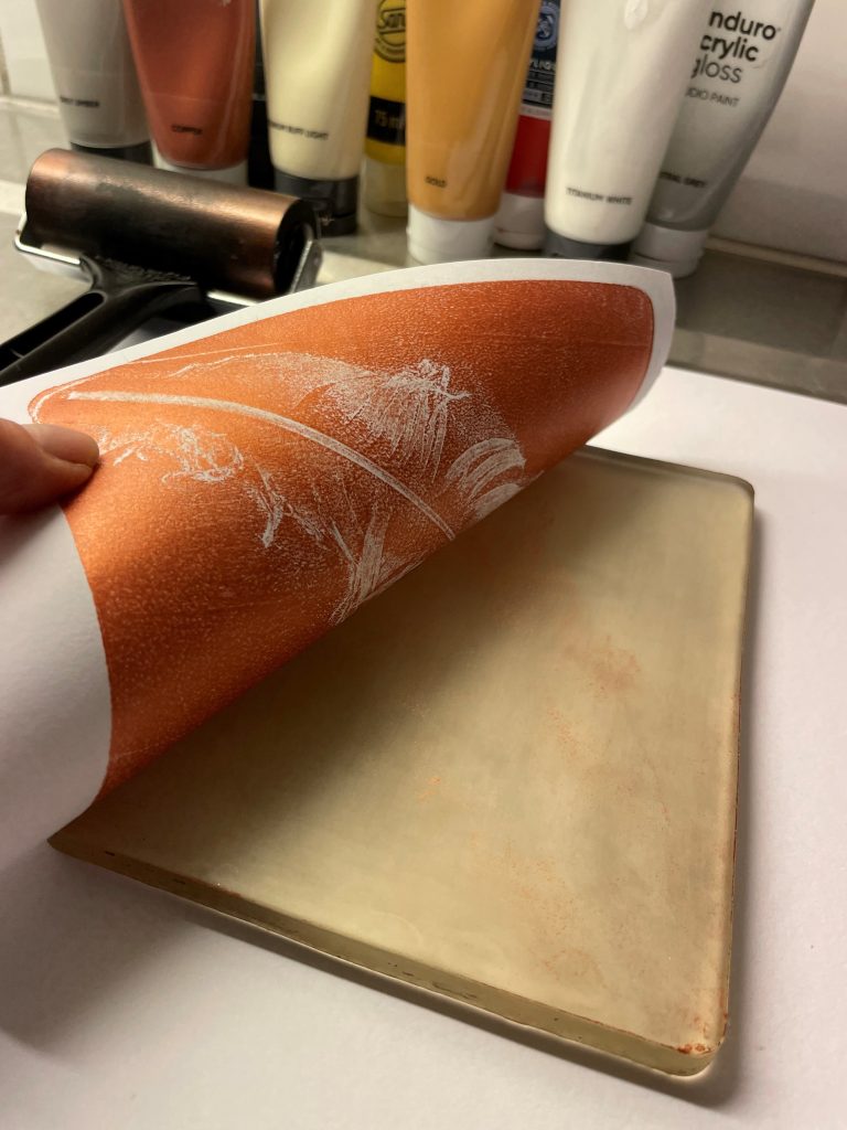 En hand lyfter upp ett papper från en infärgad tryckplåt och avslöjar ett tryck; i bakgrunden syns färgtuber.