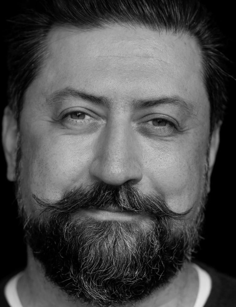 En man med tjockt skägg och mustasch som ler lätt, fotograferad i svartvitt mot en mörk bakgrund.