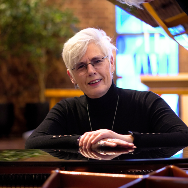En äldre kvinna med kort vitt hår lutar sig mot ett piano och ler, inomhus i ett mjukt naturligt ljus.