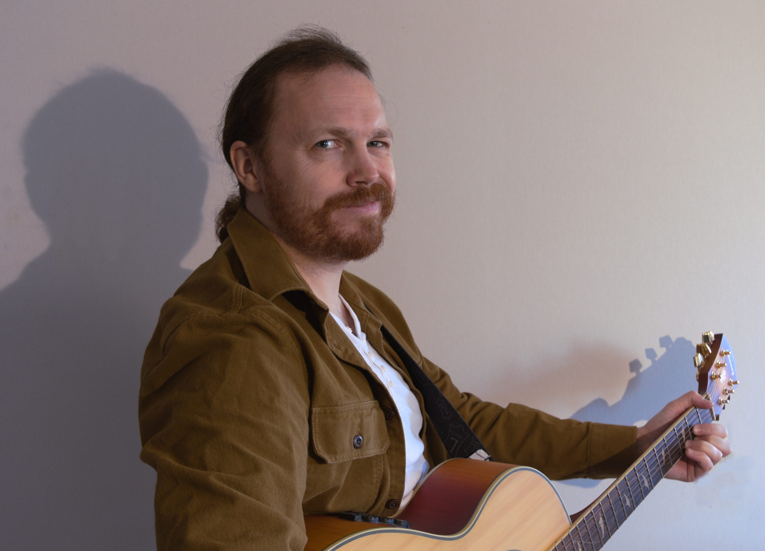 En man med brunt hår och skägg som spelar på en akustisk gitarr, klädd i en brun jacka, sittande mot en enfärgad vägg.