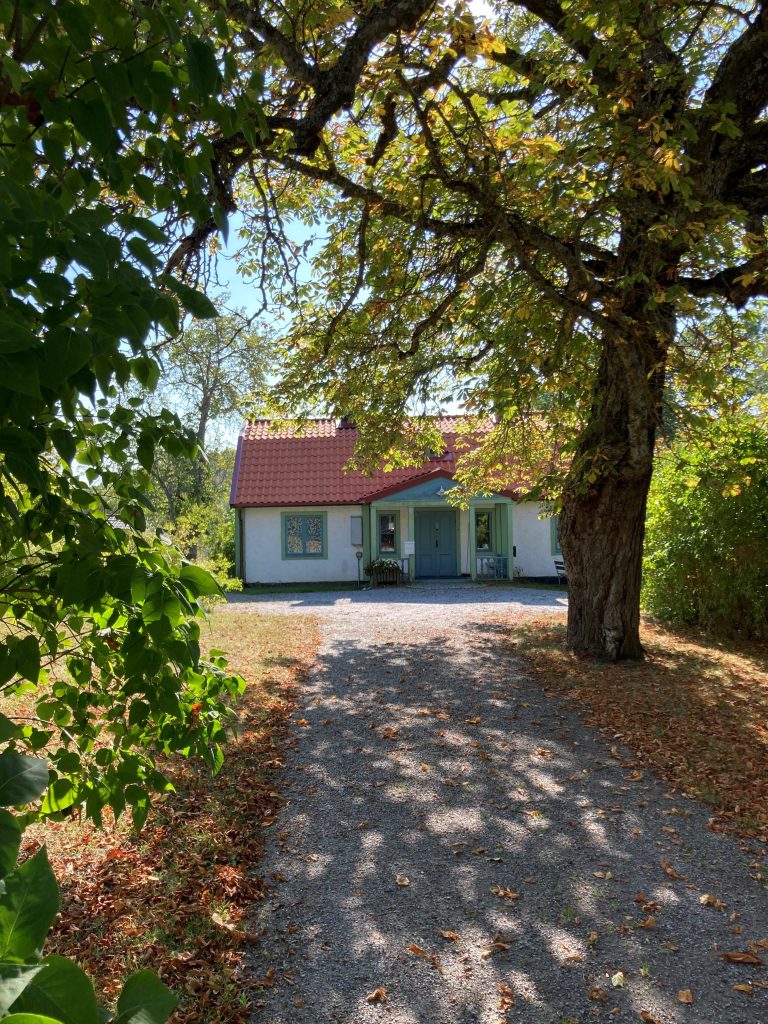 En grusgång leder fram till ett litet hus med rött tak, omgivet av stora träd och höstlöv.