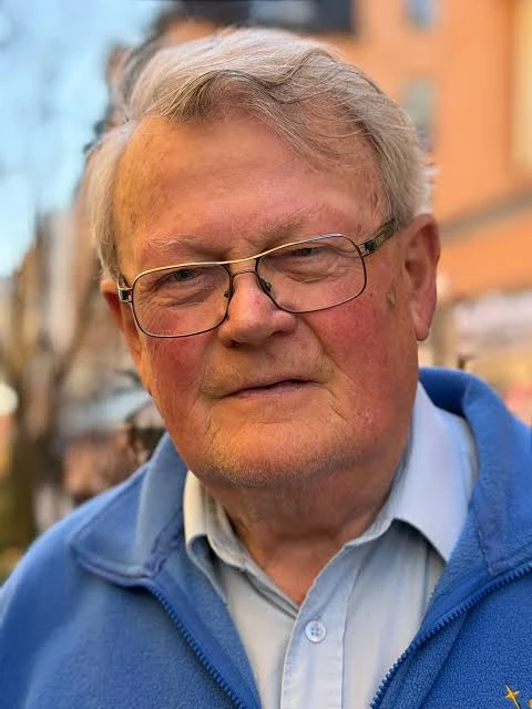 En äldre man med glasögon, grått hår och en blå jacka, som står utomhus med byggnader i bakgrunden.