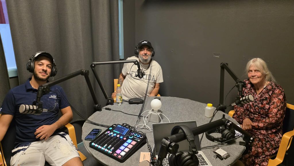 Tre personer sitter runt ett bord med mikrofoner, hörlurar och podcastutrustning i en inspelningsstudio.