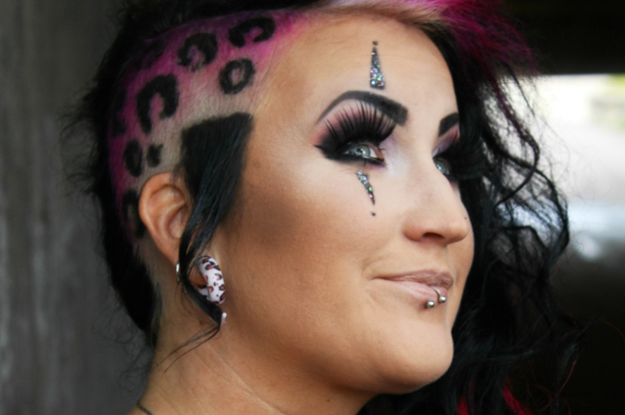 En kvinna med markant smink, rakat hår i leopardmönster, piercingar och ansiktsdekorationer av strass, som ler svagt.