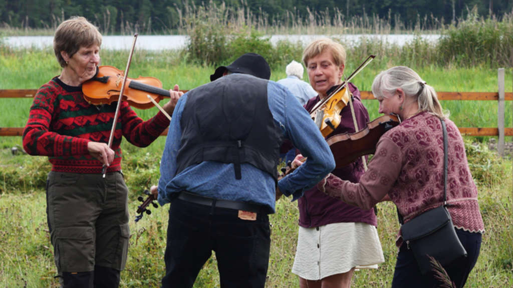Fyra personer som spelar fiol utomhus på gräset, med en sjö och träd i bakgrunden.