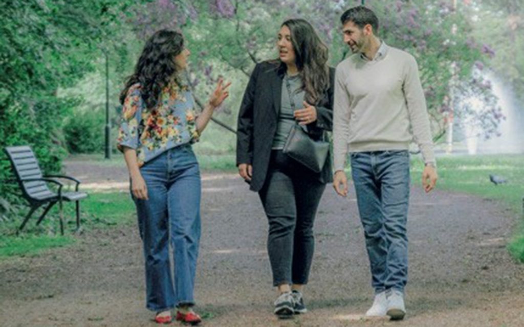 Tre personer promenerar och pratar på en trädkantad stig i en park; de verkar glada och upptagna av varandra.