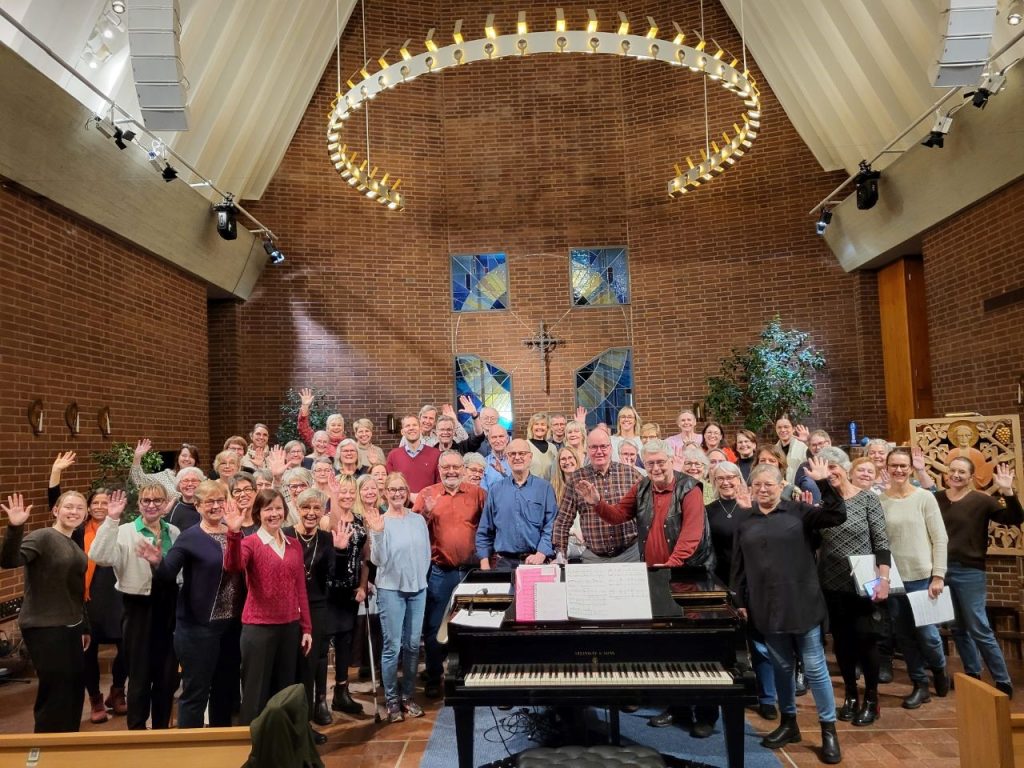 En stor körgrupp poserar och vinkar inne i en kyrka, med ett piano och noter i förgrunden.