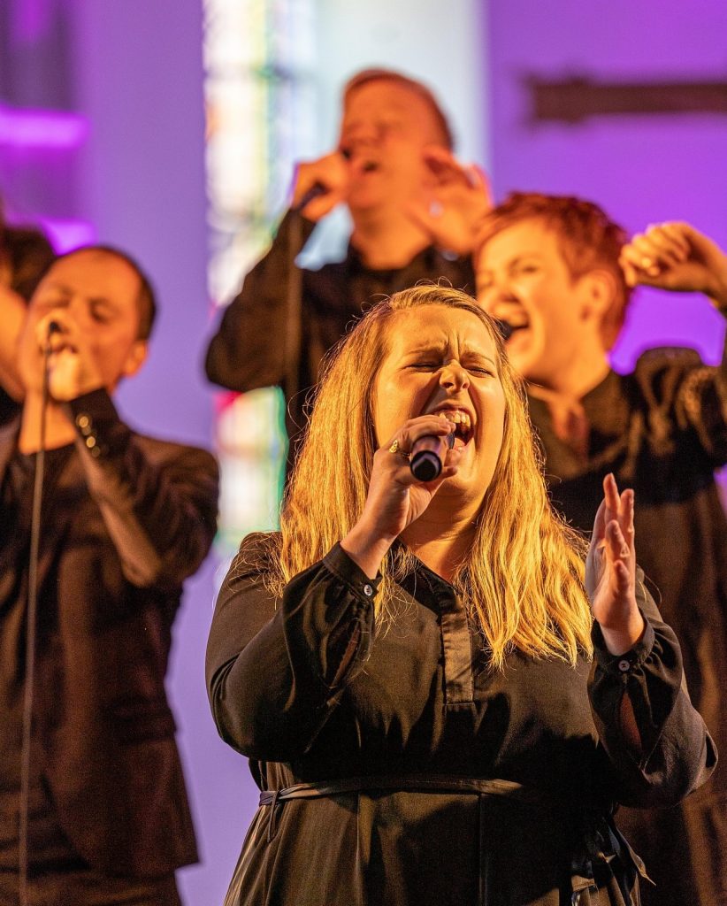 Fyra sångare uppträder med stor passion på scenen, var och en med en mikrofon i handen, under färgglada strålkastare.
