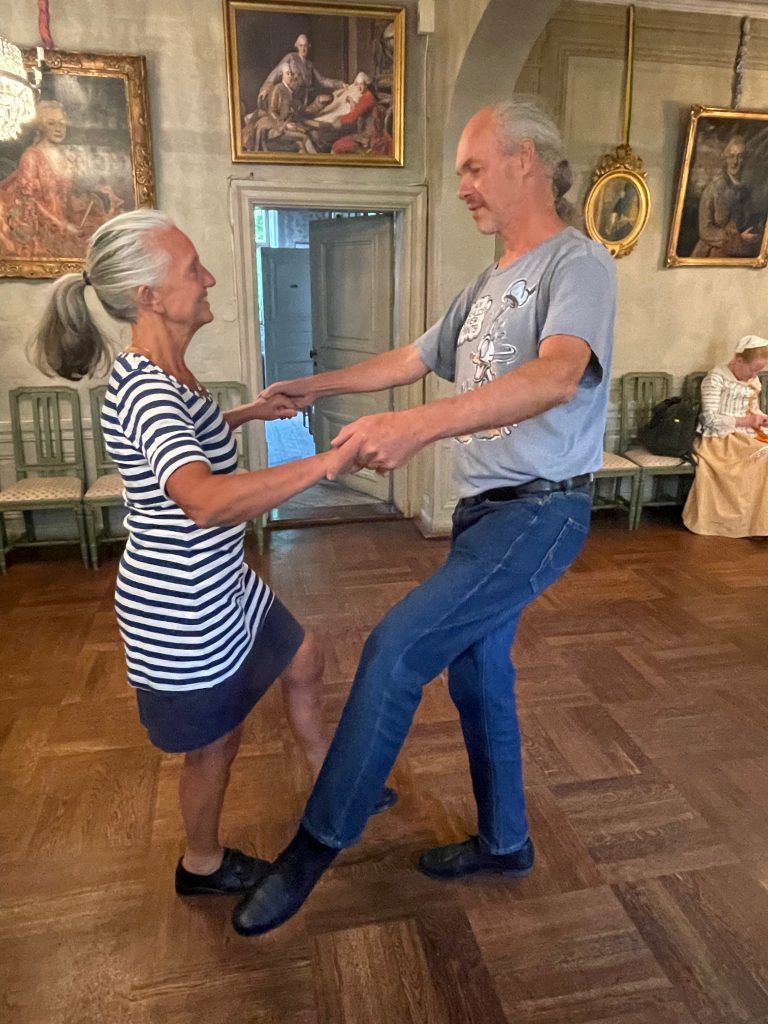 Två äldre vuxna dansar tillsammans, leende, i ett rum med trägolv och klassiska tavlor på väggarna.