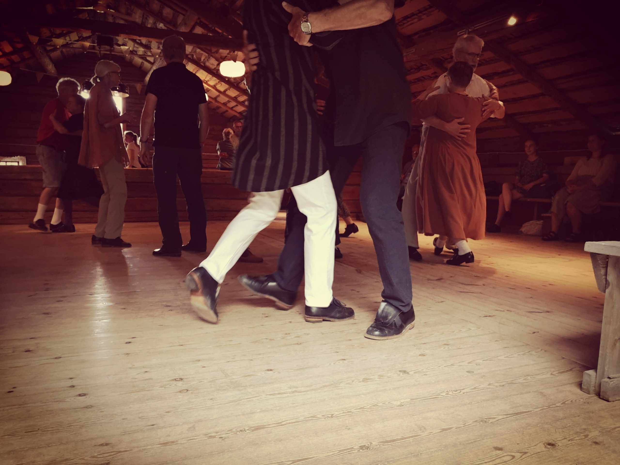 Människor som dansar i par på ett trägolv i ett rustikt rum, medan några sitter i bakgrunden.