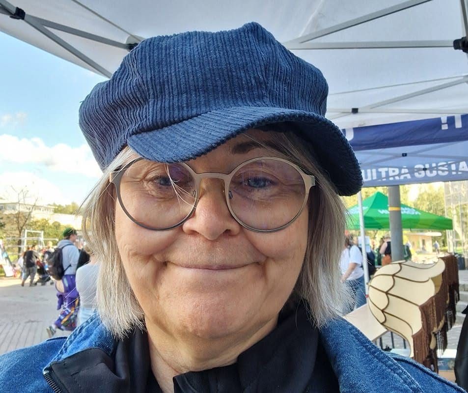 En leende person med glasögon och blå mössa under ett tält vid ett utomhusevenemang med människor i bakgrunden.