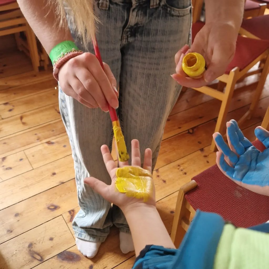 En person målar ett barns handflata gul, medan ett annat barns hand målas blå i närheten.
