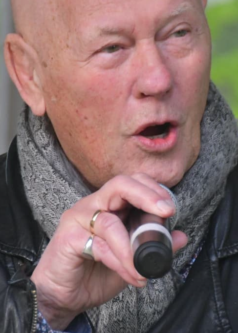 En skallig man med halsduk håller i en mikrofon och sjunger eller talar; han bär en läderjacka och en ring.