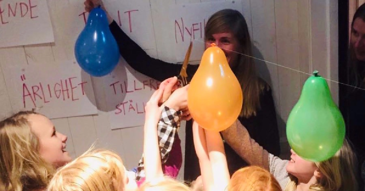 Barnen klipper av ett snöre med färgglada ballonger på en fest; på väggen bakom dem hänger affischer med texter.