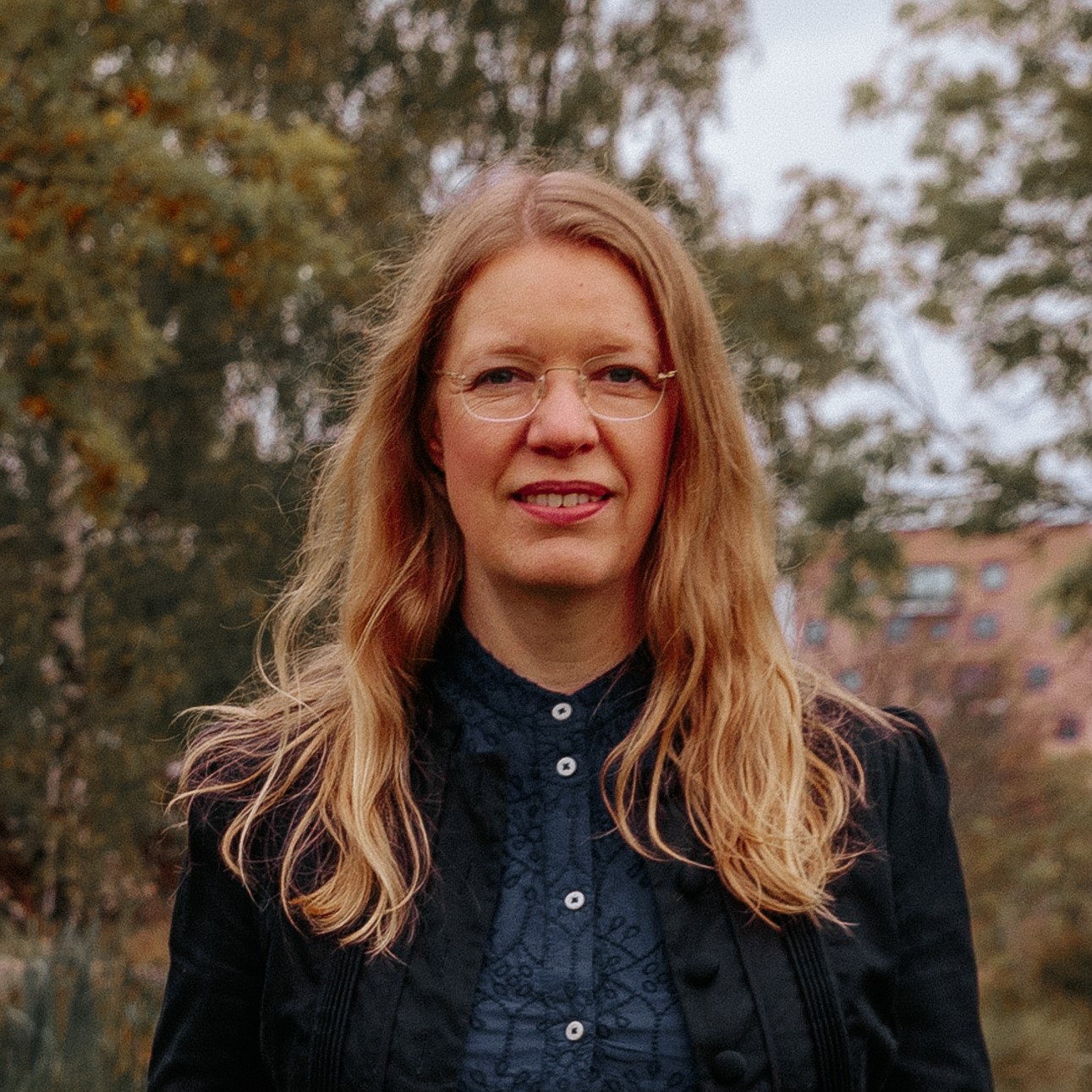 En kvinna med långt blont hår och glasögon som står utomhus med träd och byggnader i bakgrunden.