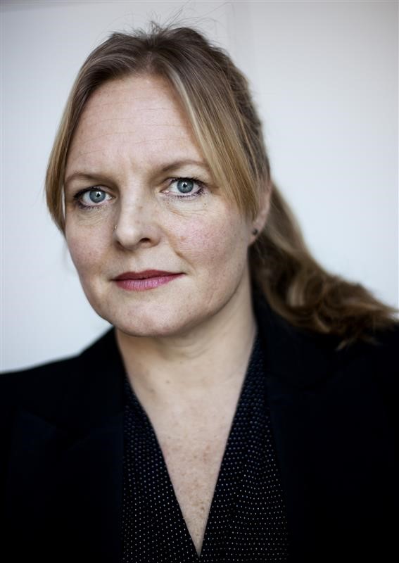 En kvinna med ljust hår i hästsvans, klädd i en svart kavaj, som tittar allvarligt in i kameran.