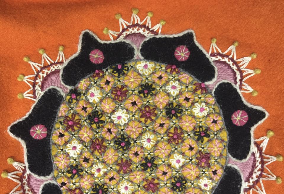 Färgglatt cirkulärt broderi med intrikata blommönster och svarta former på en orange tygbakgrund.