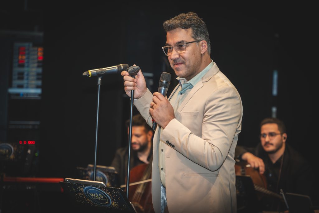 En man i beige kostym som står på scenen med en mikrofon i handen, medan musiker spelar på sina instrument i bakgrunden.
